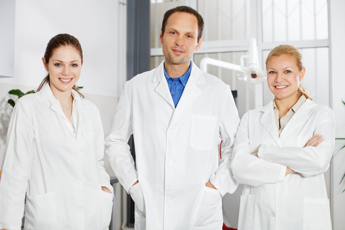 Three dental professionals in white coats posing together in a modern clinical office setting.