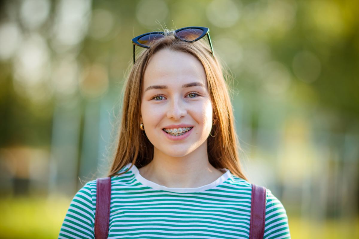 Young girl smiling outdoors wearing striped shirt and sunglasses on head, blurred green background.