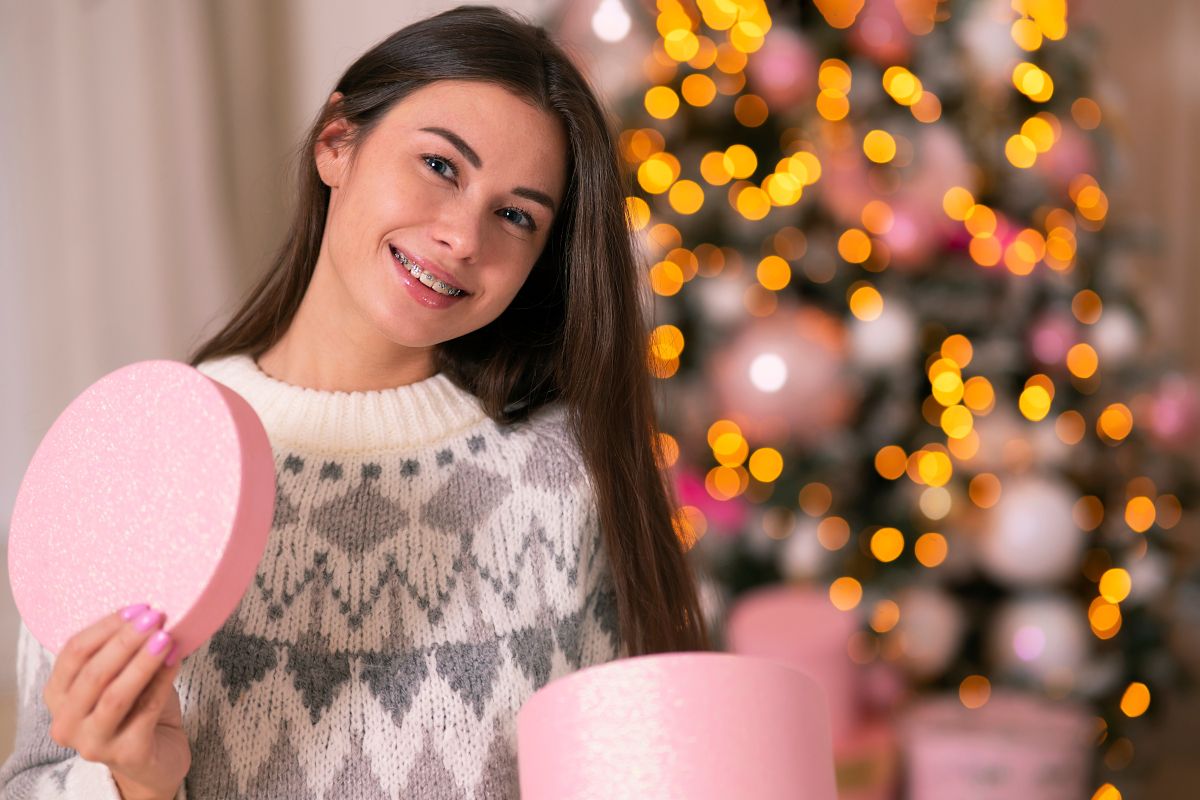 Young woman holding pink heart-shaped box, smiling at camera near decorated Christmas tree.