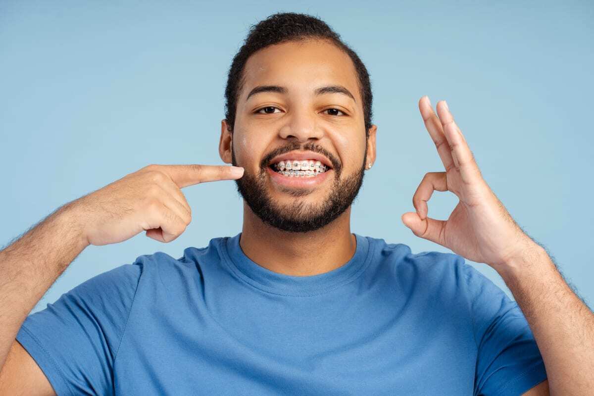 Braces in Pearl Man with metal braces smiling and pointing to his teeth against blue background.
